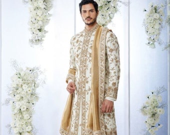 Puede incluir: Un sherwani blanco para hombre bordado con ribetes dorados y una bufanda dorada a juego. El sherwani es una prenda tradicional india que se usa para ocasiones especiales.