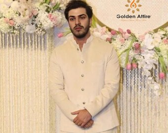 May include: A man wearing a light beige blazer with a mandarin collar and button closure. He is standing in front of a white floral backdrop with the text "Golden Attire" visible.