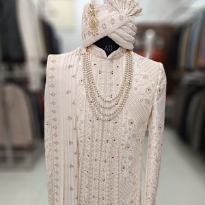 Puede incluir: Una kurta blanca bordada para hombre con una dupatta blanca a juego y un turbante blanco con detalles dorados. La kurta tiene un collar de perlas con un colgante dorado.