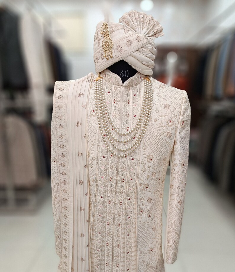 Puede incluir: Una kurta blanca bordada para hombre con una dupatta blanca a juego y un turbante blanco con detalles dorados. La kurta tiene un collar de perlas con un colgante dorado.
