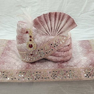 May include: A pink turban with gold and silver embellishments and a feather accent. The turban is on a matching pink fabric with a decorative border.