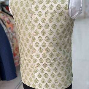 May include: A beige sleeveless waistcoat with a green geometric pattern. The waistcoat has a mandarin collar and four buttons down the front.