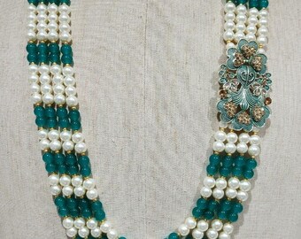 Puede incluir: Un collar de múltiples hilos con secciones alternas de perlas de imitación blancas y cuentas de color verde azulado. El collar tiene una pieza central floral decorativa de color verde azulado y dorado. El collar se exhibe en un maniquí.
