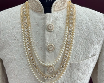 Pode incluir: Uma peça de roupa branca bordada para homem com um colar de três fios de pérolas e strass. O colar tem uma corrente dourada e pérolas brancas.