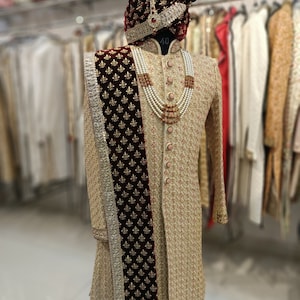 May include: A beige embroidered men's sherwani with a maroon velvet shawl and a pearl and red bead necklace. The sherwani has a mandarin collar and is worn with a maroon turban with a gold trim and a white feather.