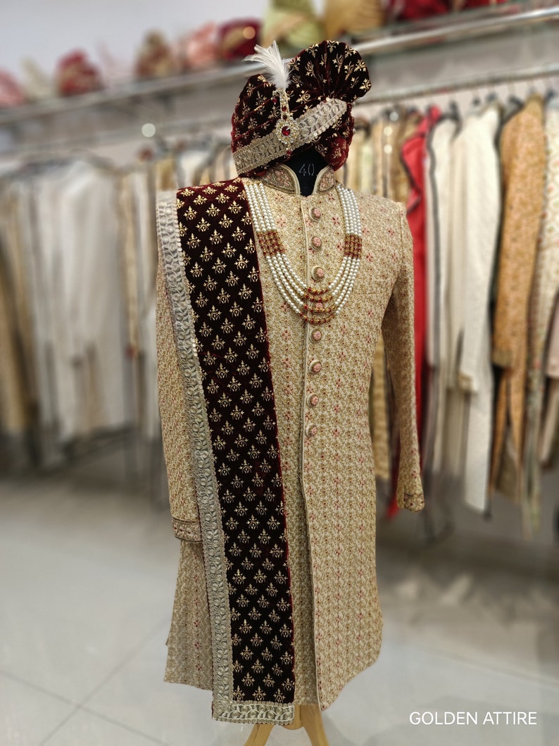 May include: A beige embroidered men's sherwani with a maroon velvet shawl and a pearl and red bead necklace. The sherwani has a mandarin collar and is worn with a maroon turban with a gold trim and a white feather.