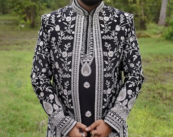 Puede incluir: Una chaqueta larga negra con un patrón floral plateado y cuello alto. La chaqueta está bordada con hilo plateado y tiene un diseño floral. La persona que lleva la chaqueta está de pie frente a un fondo verde.
