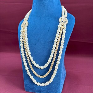 K&ouml;nnte beinhalten: Eine mehrreihige Perlenkette mit goldenen Akzenten und einem gro&szlig;en, verzierten Anh&auml;nger mit Strasssteinen. Die Kette wird auf einer blauen Samt-Puppe pr&auml;sentiert.