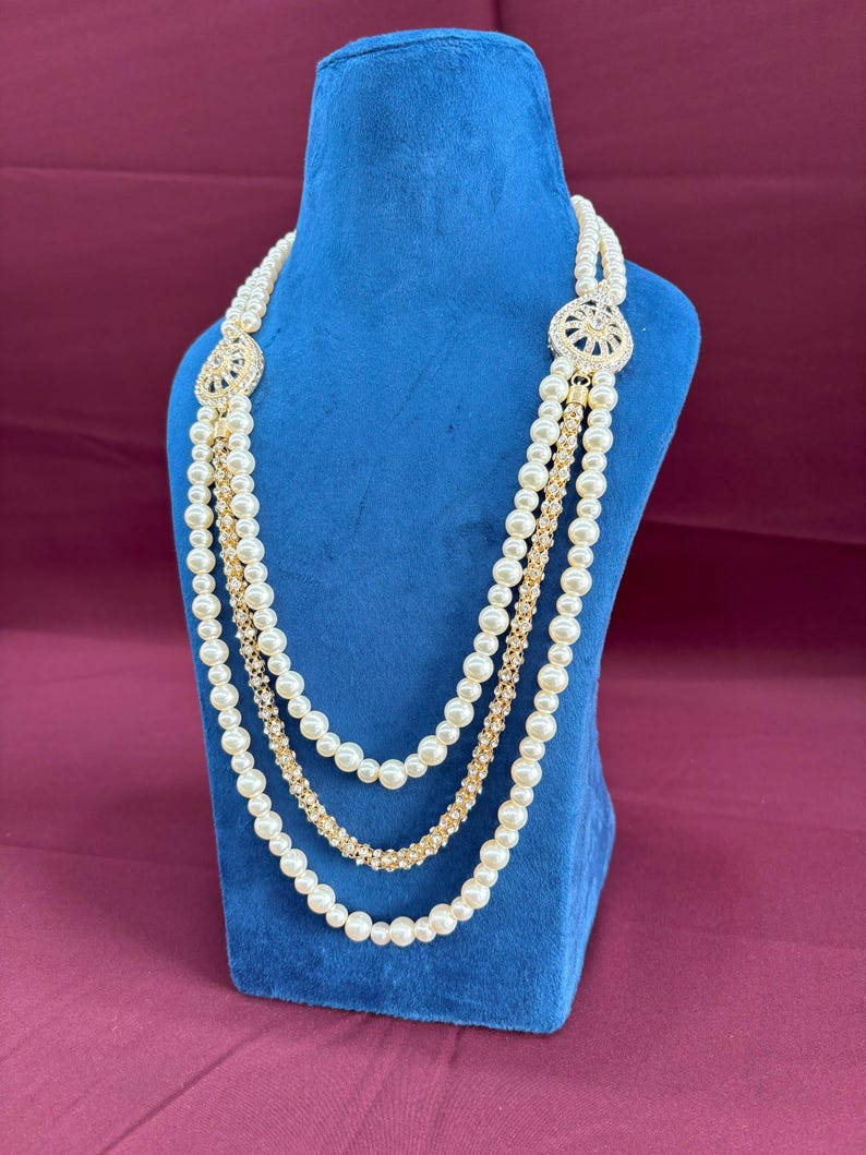 K&ouml;nnte beinhalten: Eine mehrreihige Perlenkette mit goldenen Akzenten und einem gro&szlig;en, verzierten Anh&auml;nger mit Strasssteinen. Die Kette wird auf einer blauen Samt-Puppe pr&auml;sentiert.