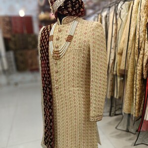 May include: A gold embroidered men's sherwani with a maroon velvet shawl and a pearl necklace. The sherwani has a mandarin collar and is worn on a mannequin. The text "GOLDEN ATTIRE" is visible in the bottom right corner of the image.