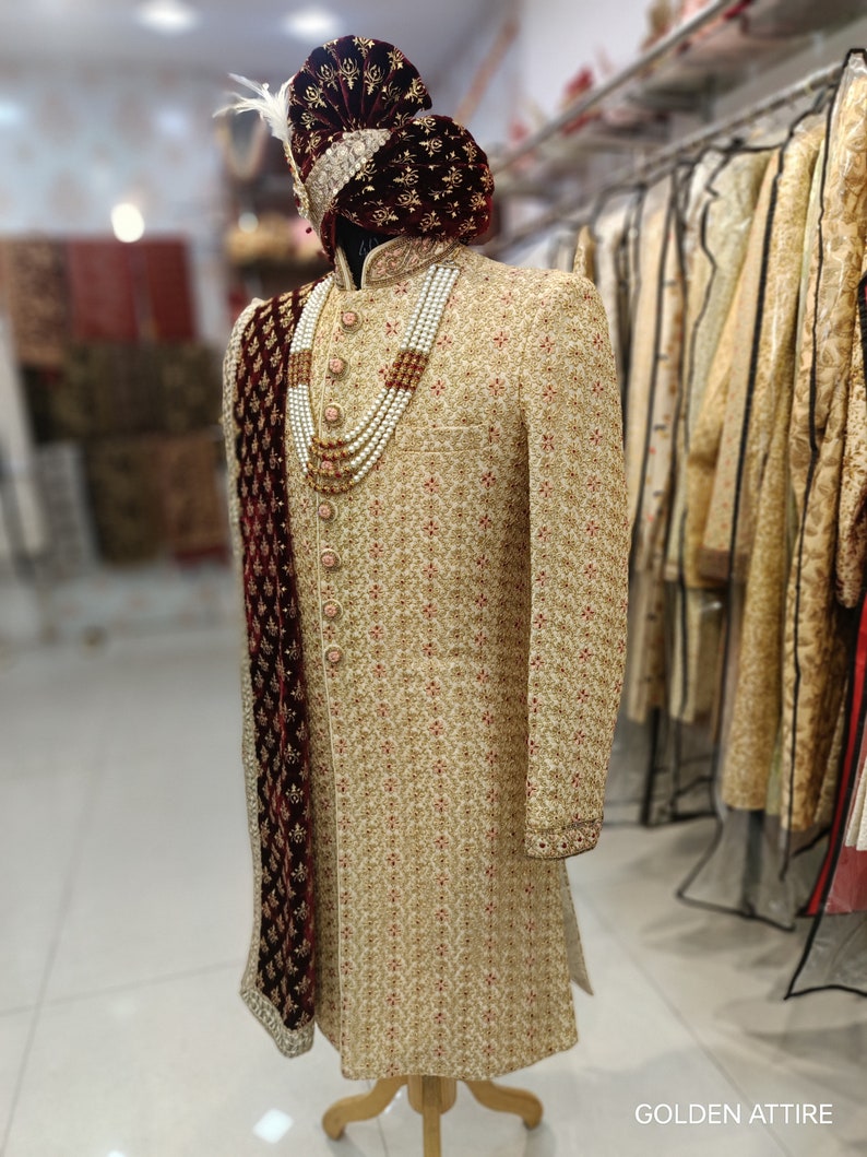 May include: A gold embroidered men's sherwani with a maroon velvet shawl and a pearl necklace. The sherwani has a mandarin collar and is worn on a mannequin. The text "GOLDEN ATTIRE" is visible in the bottom right corner of the image.