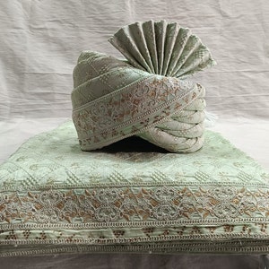 May include: A light green turban with gold and white embroidery. The turban is folded into a fan shape and is sitting on a matching light green fabric with gold and white embroidery.