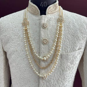 K&ouml;nnte beinhalten: Eine wei&szlig;e bestickte Herrenjacke mit goldener Einfassung und einer dreireihigen Perlenkette mit goldenen Akzenten.