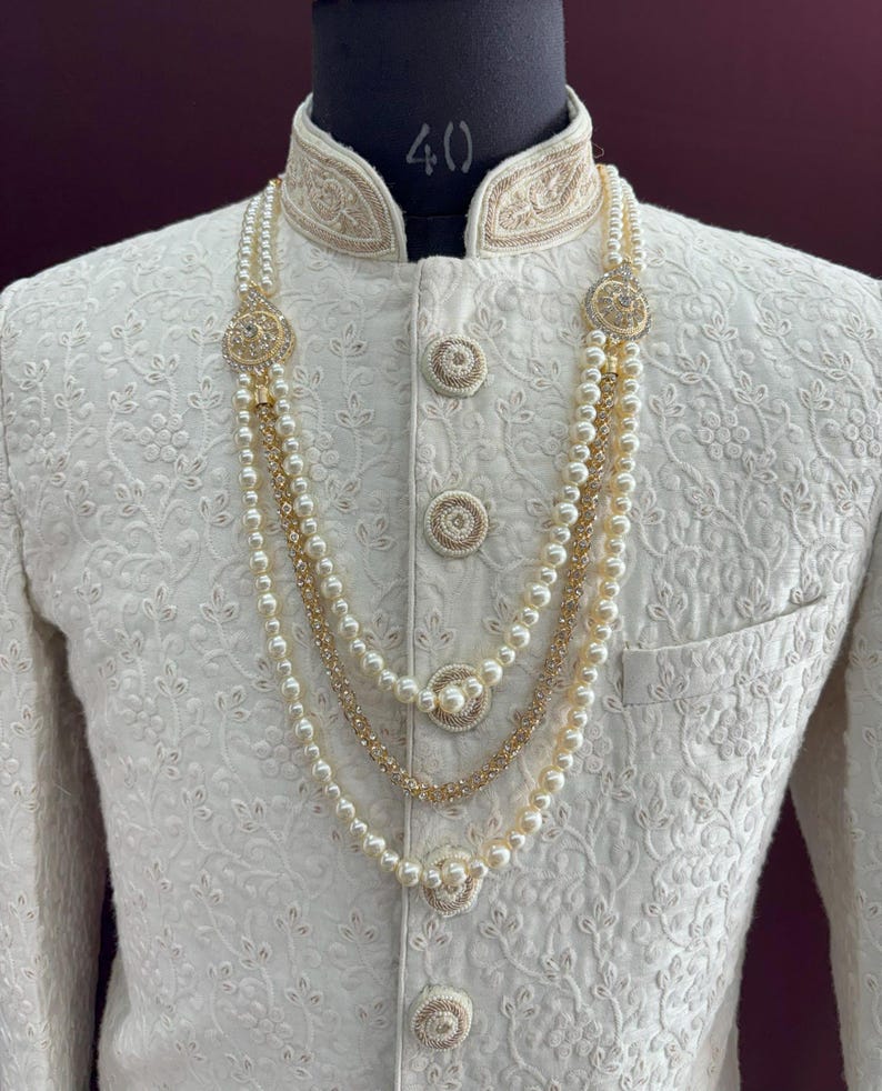 K&ouml;nnte beinhalten: Eine wei&szlig;e bestickte Herrenjacke mit goldener Einfassung und einer dreireihigen Perlenkette mit goldenen Akzenten.