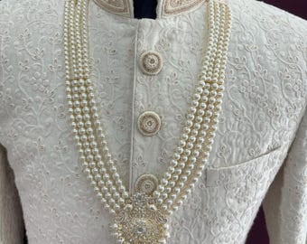 Può includere: Un capo di abbigliamento bianco ricamato per uomo con una collana di perle e un grande ciondolo ornato. Il ciondolo è in oro e ha una pietra centrale che sembra un diamante.