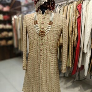 May include: A gold embroidered men's sherwani with a maroon velvet turban and a pearl and gold necklace. The sherwani has a mandarin collar and button closure. The text "GOLDEN ATTIRE" is visible in the bottom right corner of the image.