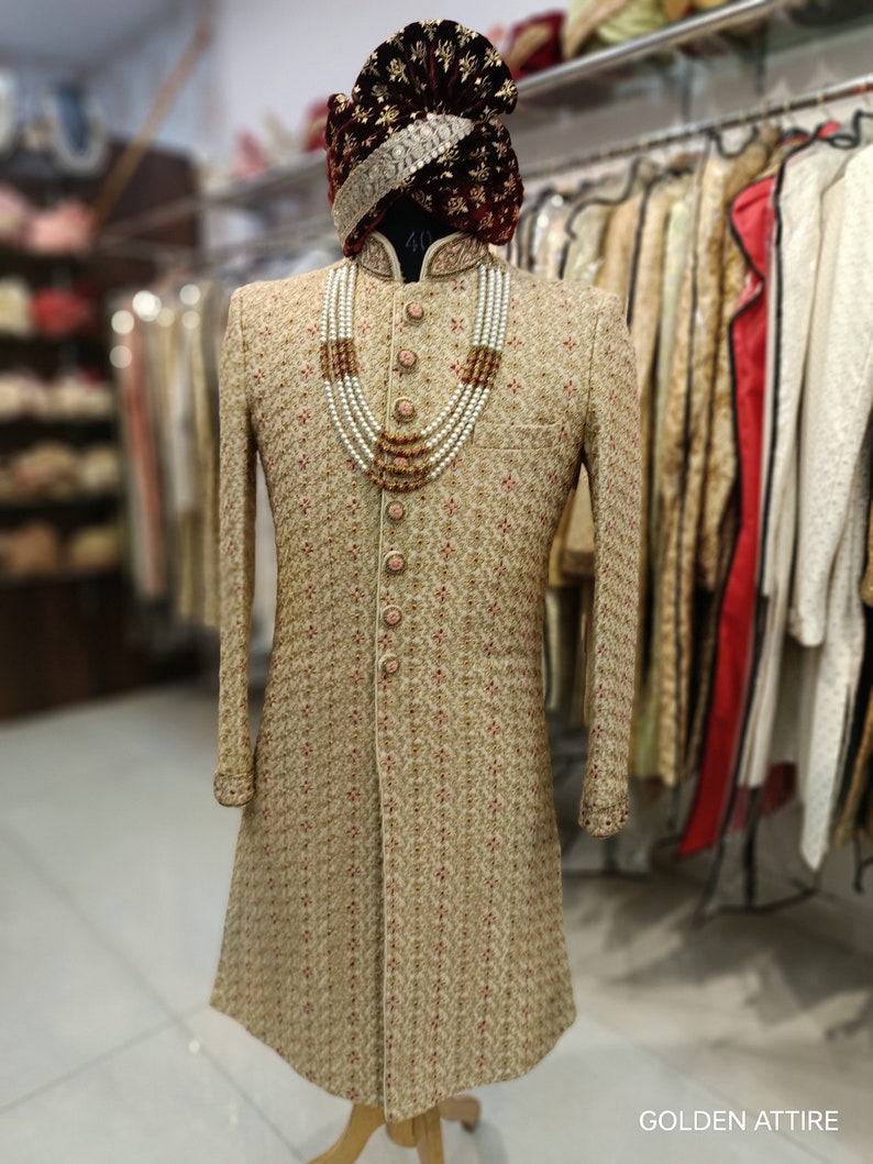 May include: A gold embroidered men's sherwani with a maroon velvet turban and a pearl and gold necklace. The sherwani has a mandarin collar and button closure. The text "GOLDEN ATTIRE" is visible in the bottom right corner of the image.