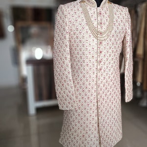 May include: Cream-colored, long-sleeved embroidered coat with a mandarin collar, displayed on a wooden mannequin. The coat features a repeating pattern of red and cream-colored floral designs. A layered pearl necklace is draped around the neck.