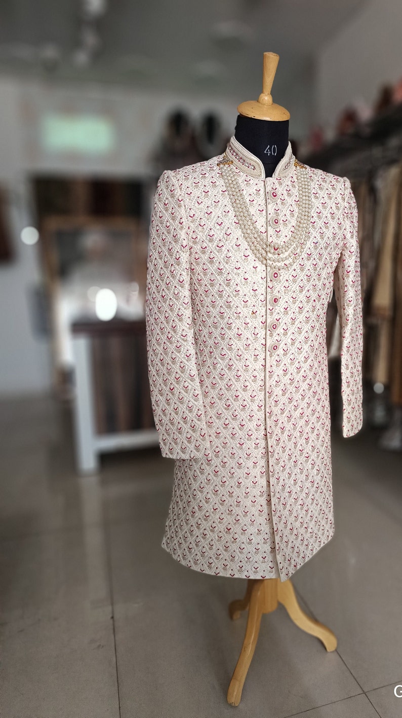 May include: Cream-colored, long-sleeved embroidered coat with a mandarin collar, displayed on a wooden mannequin. The coat features a repeating pattern of red and cream-colored floral designs. A layered pearl necklace is draped around the neck.