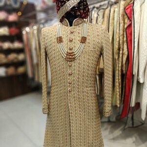 May include: A gold patterned sherwani with a maroon velvet turban and a pearl and orange bead necklace. The sherwani has a mandarin collar and a button closure. The turban has a gold trim. The necklace is layered and has a decorative pendant. The text "GOLDEN ATTIRE" is visible at the bottom of the image.
