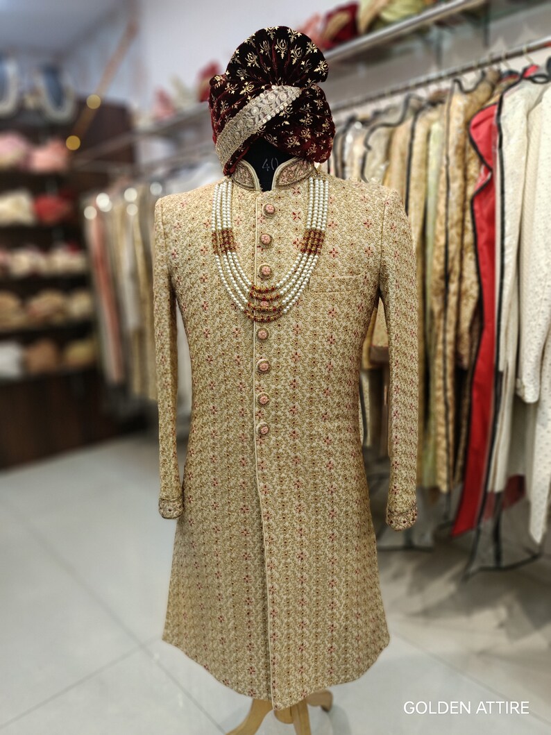 May include: A gold patterned sherwani with a maroon velvet turban and a pearl and orange bead necklace. The sherwani has a mandarin collar and a button closure. The turban has a gold trim. The necklace is layered and has a decorative pendant. The text "GOLDEN ATTIRE" is visible at the bottom of the image.
