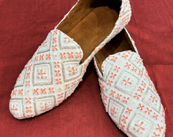Pode incluir: Um par de mocassins brancos bordados com um padrão floral rosa. Os mocassins têm uma ponta pontiaguda e um design de deslizamento.