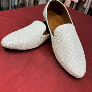 Pode incluir: Um par de sapatos de mocassim brancos bordados com um padr&atilde;o texturizado. Os mocassins est&atilde;o sobre uma superf&iacute;cie vermelha.