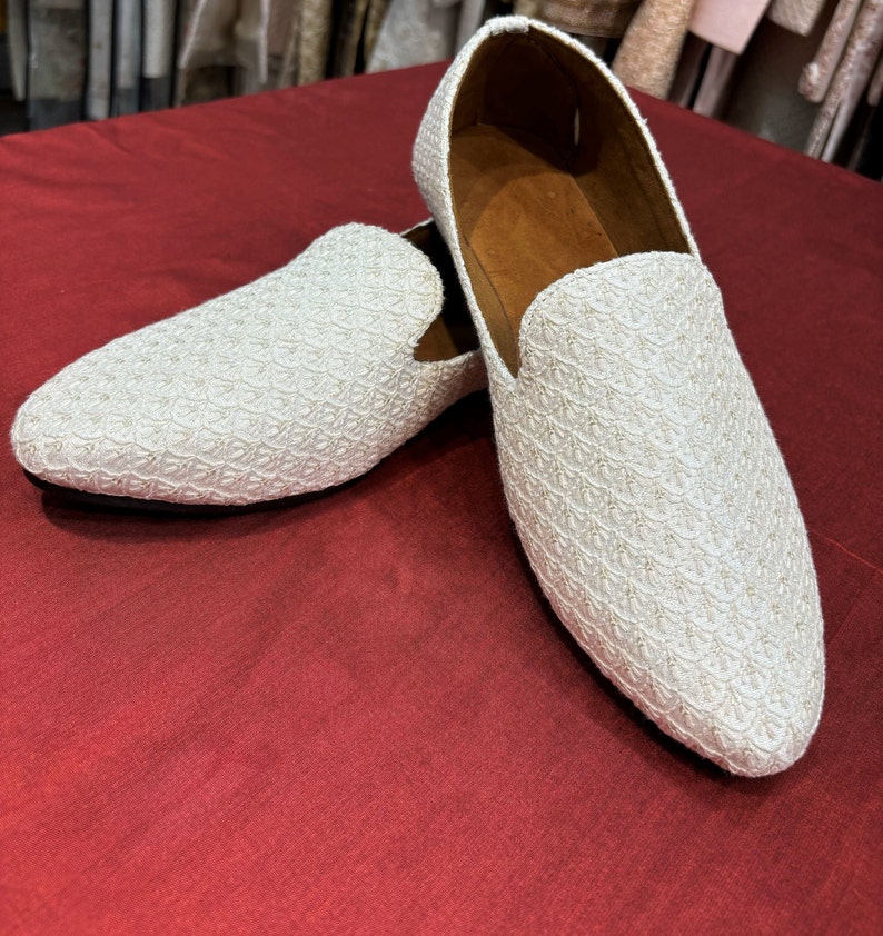 Pode incluir: Um par de sapatos de mocassim brancos bordados com um padr&atilde;o texturizado. Os mocassins est&atilde;o sobre uma superf&iacute;cie vermelha.