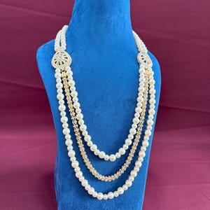 K&ouml;nnte beinhalten: Eine dreireihige Perlenkette mit goldenen Akzenten und einem gro&szlig;en, verzierten Anh&auml;nger mit Strasssteinen. Die Kette wird auf einem blauen Samtst&auml;nder pr&auml;sentiert.