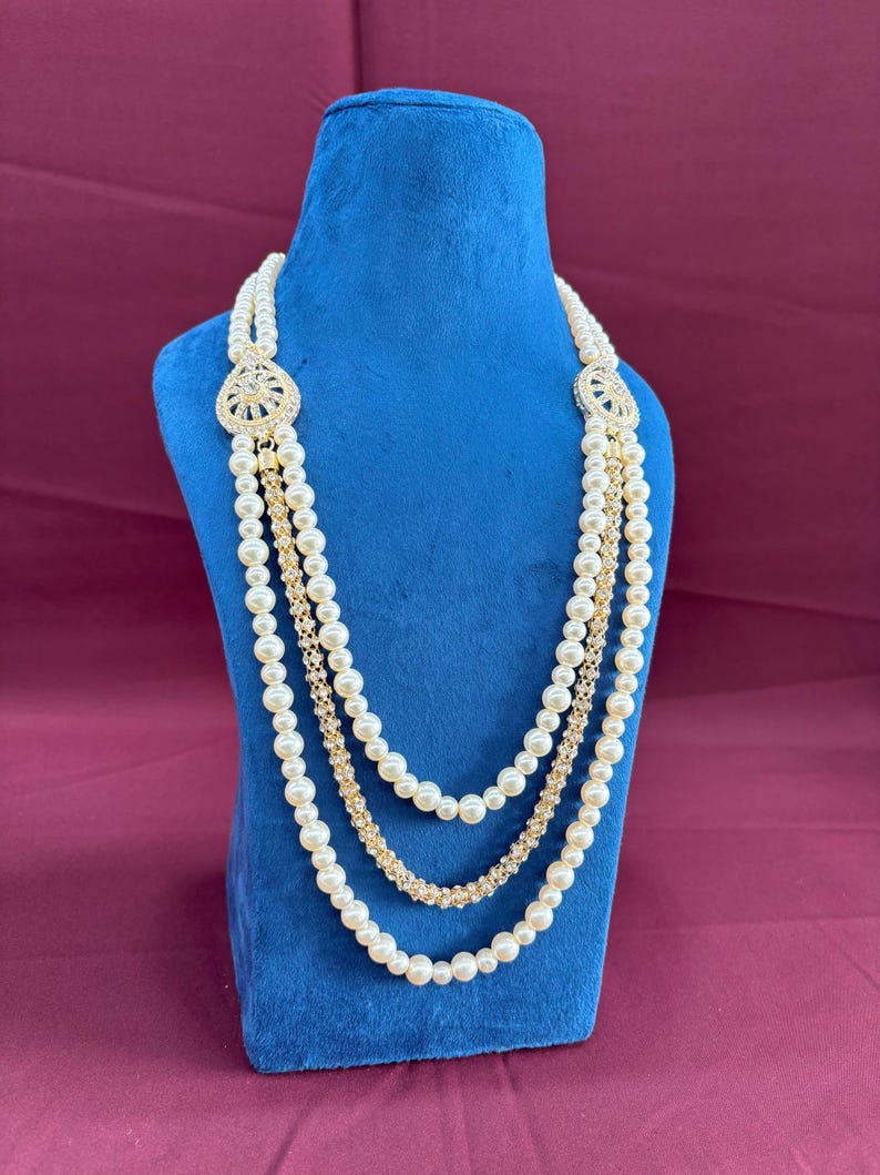 K&ouml;nnte beinhalten: Eine dreireihige Perlenkette mit goldenen Akzenten und einem gro&szlig;en, verzierten Anh&auml;nger mit Strasssteinen. Die Kette wird auf einem blauen Samtst&auml;nder pr&auml;sentiert.