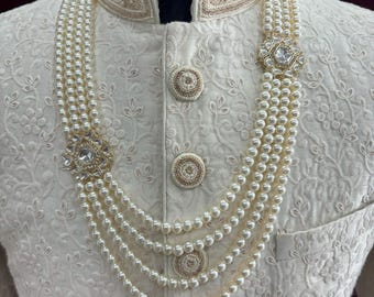 May include: A white pearl necklace with four strands and two gold and clear rhinestone embellishments. The necklace is worn on a white garment with gold embroidery.