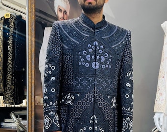 Peut inclure: Un sherwani brodé bleu marine foncé avec des motifs floraux et géométriques complexes. Le long manteau formel présente un col mandarin et est orné d'accents blancs et argentés. Le vêtement est présenté sur une personne dans un cadre de magasin.