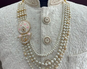Può includere: Una collana di perle bianche con un ciondolo dorato e bianco. La collana è a strati e presenta un design decorativo. La collana è indossata su un capo bianco con ricami dorati.
