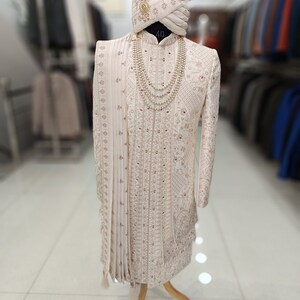 Puede incluir: Un sherwani blanco bordado para hombre con un turbante blanco a juego y un collar de perlas. El sherwani tiene un ribete dorado y un dise&ntilde;o rojo y dorado. El turbante tiene una borla dorada. La imagen est&aacute; etiquetada como "GOLDEN ATTIRE".