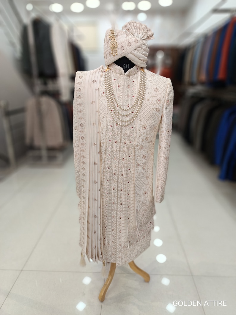 Puede incluir: Un sherwani blanco bordado para hombre con un turbante blanco a juego y un collar de perlas. El sherwani tiene un ribete dorado y un dise&ntilde;o rojo y dorado. El turbante tiene una borla dorada. La imagen est&aacute; etiquetada como "GOLDEN ATTIRE".