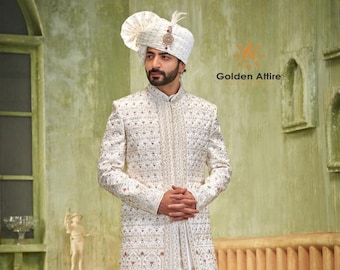 Puede incluir: Un sherwani blanco bordado con un turbante blanco. El sherwani tiene un diseño largo y fluido y está decorado con bordados intrincados. El turbante está adornado con una pluma blanca.