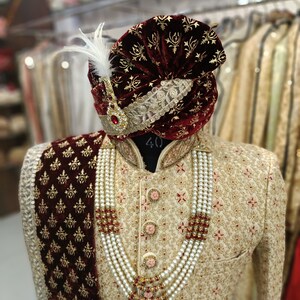May include: A gold and cream patterned men's sherwani with a maroon velvet shawl and a maroon velvet turban with gold embroidery. The sherwani has a pearl necklace with gold accents. The text "GOLDEN ATTIRE" is visible in the bottom right corner of the image.