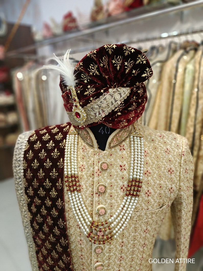 May include: A gold and cream patterned men's sherwani with a maroon velvet shawl and a maroon velvet turban with gold embroidery. The sherwani has a pearl necklace with gold accents. The text "GOLDEN ATTIRE" is visible in the bottom right corner of the image.