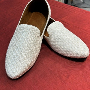 Pode incluir: Um par de mocassins brancos bordados com bico fino. Os mocassins t&ecirc;m um padr&atilde;o sutil e s&atilde;o feitos de um tecido macio e texturizado.