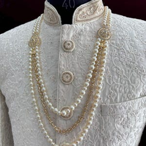 K&ouml;nnte beinhalten: Eine wei&szlig;e Perlen- und Goldkette mit dekorativen runden Akzenten. Die Kette ist mit drei Reihen Perlen und einer Goldkette mit kleinen funkelnden Steinen geschichtet.