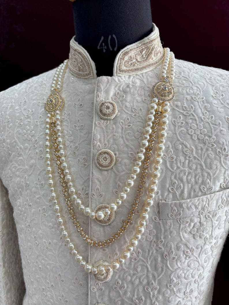 K&ouml;nnte beinhalten: Eine wei&szlig;e Perlen- und Goldkette mit dekorativen runden Akzenten. Die Kette ist mit drei Reihen Perlen und einer Goldkette mit kleinen funkelnden Steinen geschichtet.