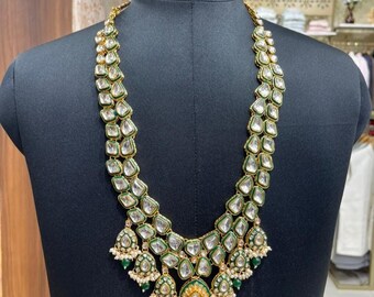 Puede incluir: Un collar dorado con detalles transparentes, verde esmeralda y blanco. El collar presenta un diseño en capas con múltiples colgantes de piedra transparente y detalles de cuentas verdes y blancas. El collar se exhibe en un maniquí negro.
