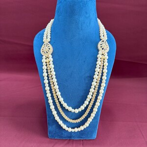 K&ouml;nnte beinhalten: Eine mehrreihige Perlenkette mit goldenen Akzenten und zwei gro&szlig;en, verzierten, juwelenbesetzten Anh&auml;ngern. Die Kette wird auf einer blauen Samt-Puppe pr&auml;sentiert.