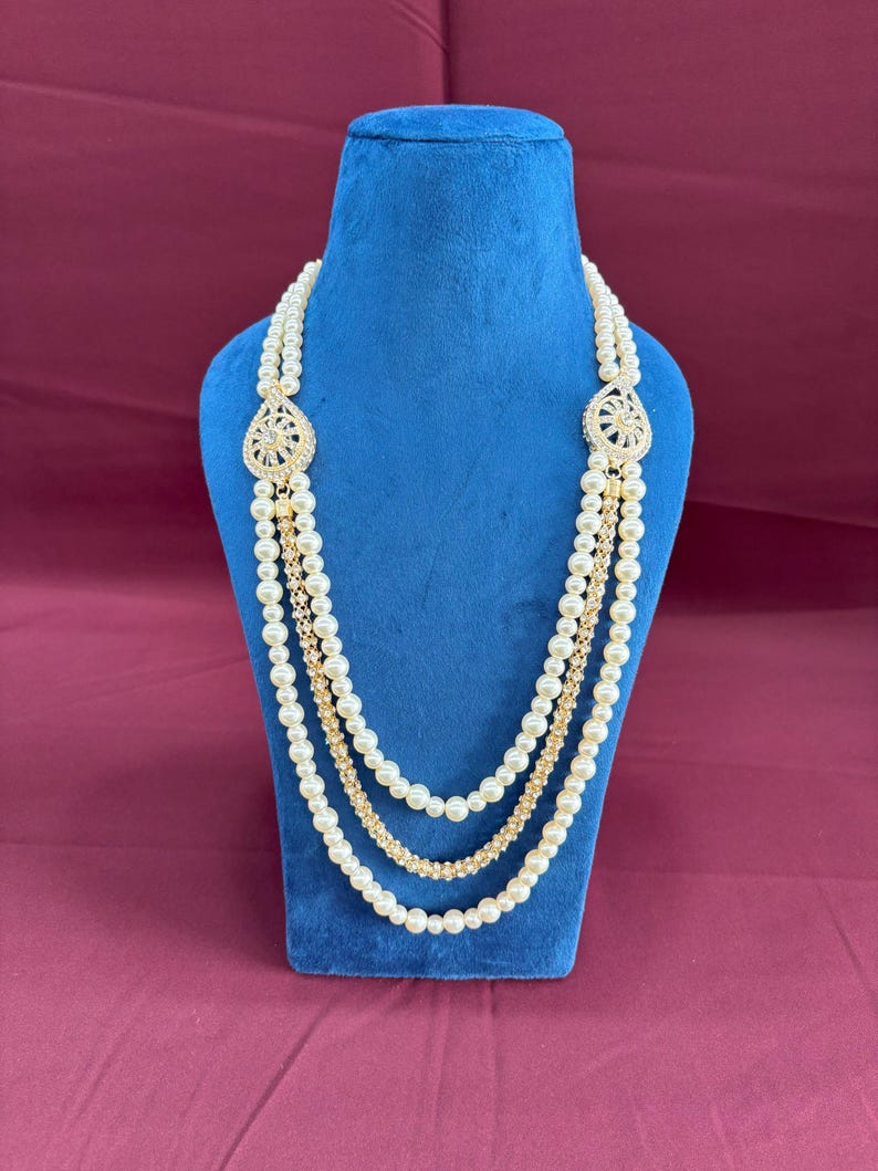 K&ouml;nnte beinhalten: Eine mehrreihige Perlenkette mit goldenen Akzenten und zwei gro&szlig;en, verzierten, juwelenbesetzten Anh&auml;ngern. Die Kette wird auf einer blauen Samt-Puppe pr&auml;sentiert.