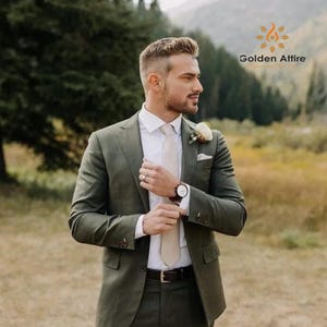 Op de afbeelding: Een man in een olijfgroen pak, wit overhemd en beige stropdas, met een bloem op de revers. Het horloge heeft een bruine leren band. De tekst "Golden Attire" staat rechtsboven. De achtergrond is een veld en bergen.