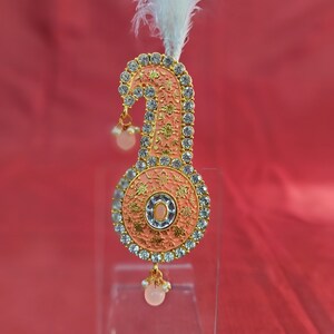 May include: A pink and gold ornate headpiece with white feathers and clear rhinestones. The headpiece has a round, decorative center with a small, pink mirror.