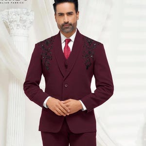 Puede incluir: Un traje de tres piezas de color granate con detalles florales bordados en negro en la solapa. El traje tiene un cierre de botón único y una camisa blanca con una corbata roja.