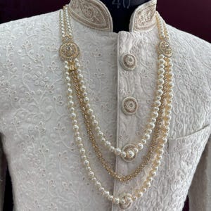 K&ouml;nnte beinhalten: Ein wei&szlig;es besticktes Herrenkleidungsst&uuml;ck mit einem goldenen Kragenbesatz und einer mehrreihigen Perlenkette mit goldenen Akzenten und einem 4( )-Etikett.