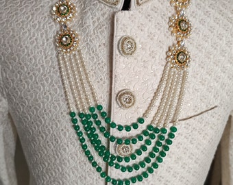 Puede incluir: Una prenda blanca para hombre con un collar de cuentas verdes y blancas. El collar tiene varias hileras de cuentas verdes y perlas blancas. La prenda tiene un cuello alto y está decorada con bordados blancos.
