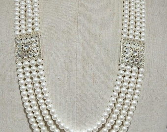 Può includere: Collana di perle a più fili con chiusure quadrate argentate adornate con cristalli trasparenti. La collana è esposta su un manichino, mettendo in risalto il suo design a strati e l'estetica elegante.